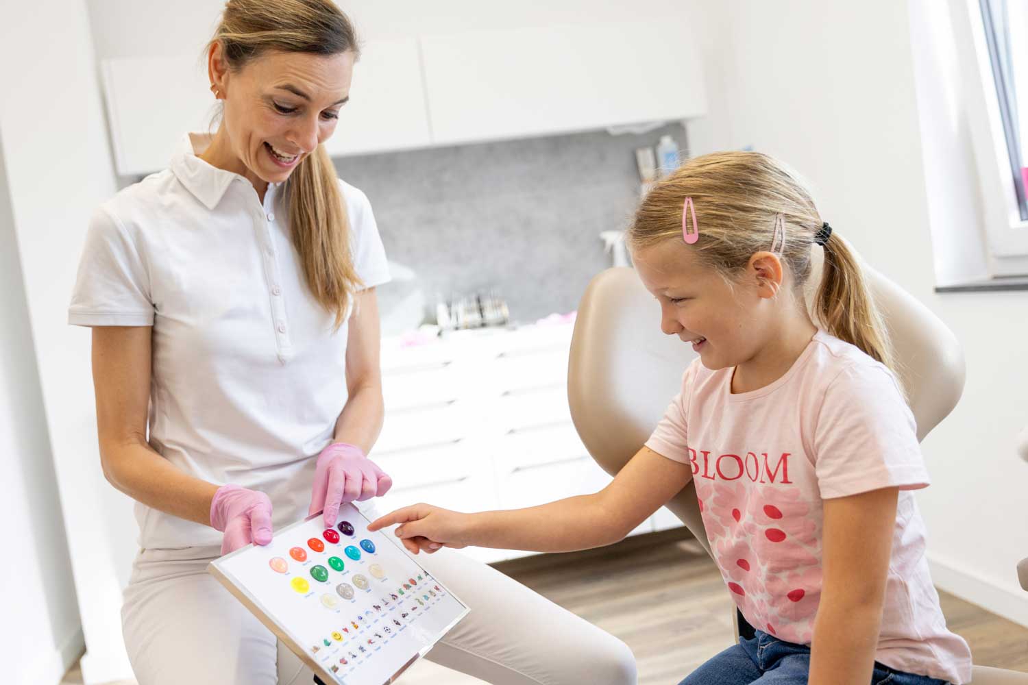 Kieferorthopädin Dr. Schumacher wählt gemeinsam mit einem Mädchen im rosa Shirt Farben für ihre Zahnspange in der Praxis Kieferorthopädie Seelscheid in Neunkirchen-Seelscheid aus.