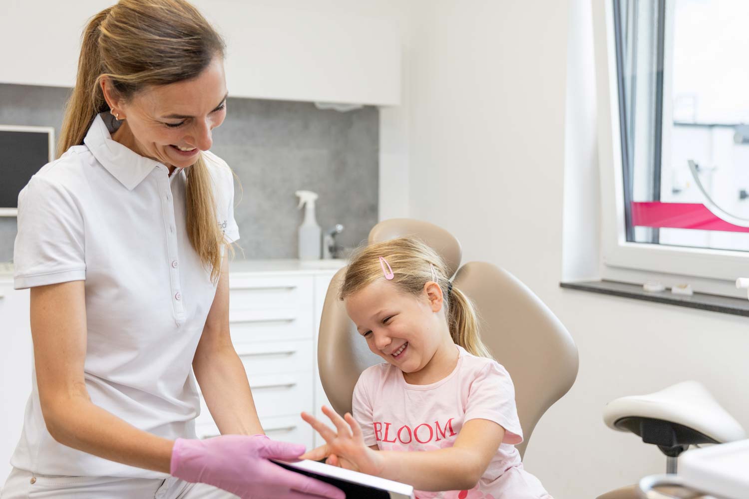 Kieferorthopädin Dr. Schumacher zeigt einem fröhlich lachenden Mädchen auf dem Behandlungsstuhl ein Tablet in der Praxis Kieferorthopädie Seelscheid in Neunkirchen-Seelscheid.