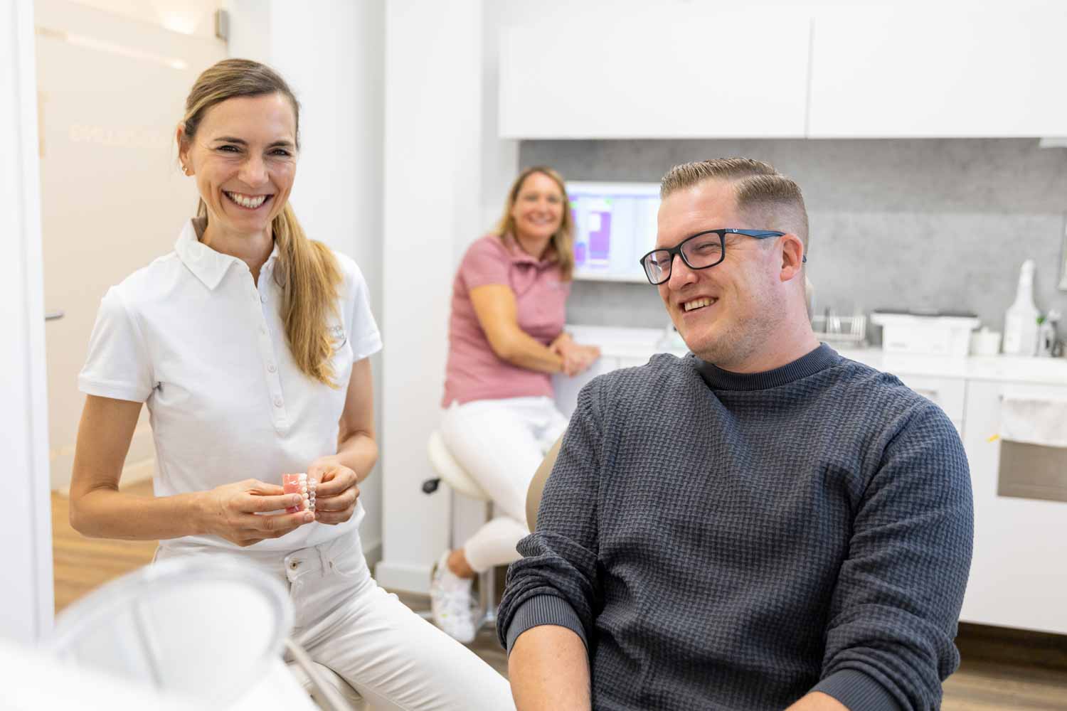 Kieferorthopädin Dr. Schumacher hält ein Gebissmodell und berät lächelnd einen erwachsenen Patienten mit Brille auf dem Behandlungsstuhl der Kieferorthopädie Seelscheid in Neunkirchen-Seelscheid.