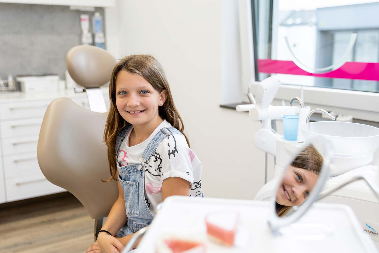 Junge Patientin blickt lächelnd in einen Handspiegel auf dem Behandlungsstuhl der Kieferorthopädie Seelscheid in Neunkirchen-Seelscheid.