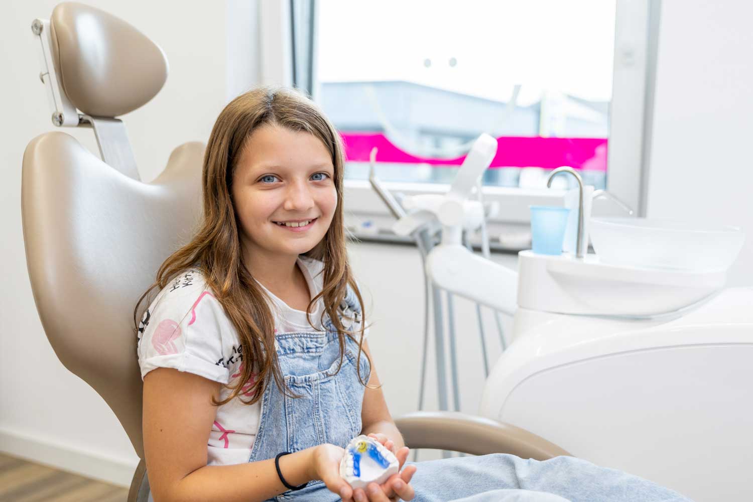 Junge Patientin hält stolz ihr Gipsmodell mit herausnehmbarer Zahnspange auf dem Behandlungsstuhl der Kieferorthopädie Seelscheid in Neunkirchen-Seelscheid.