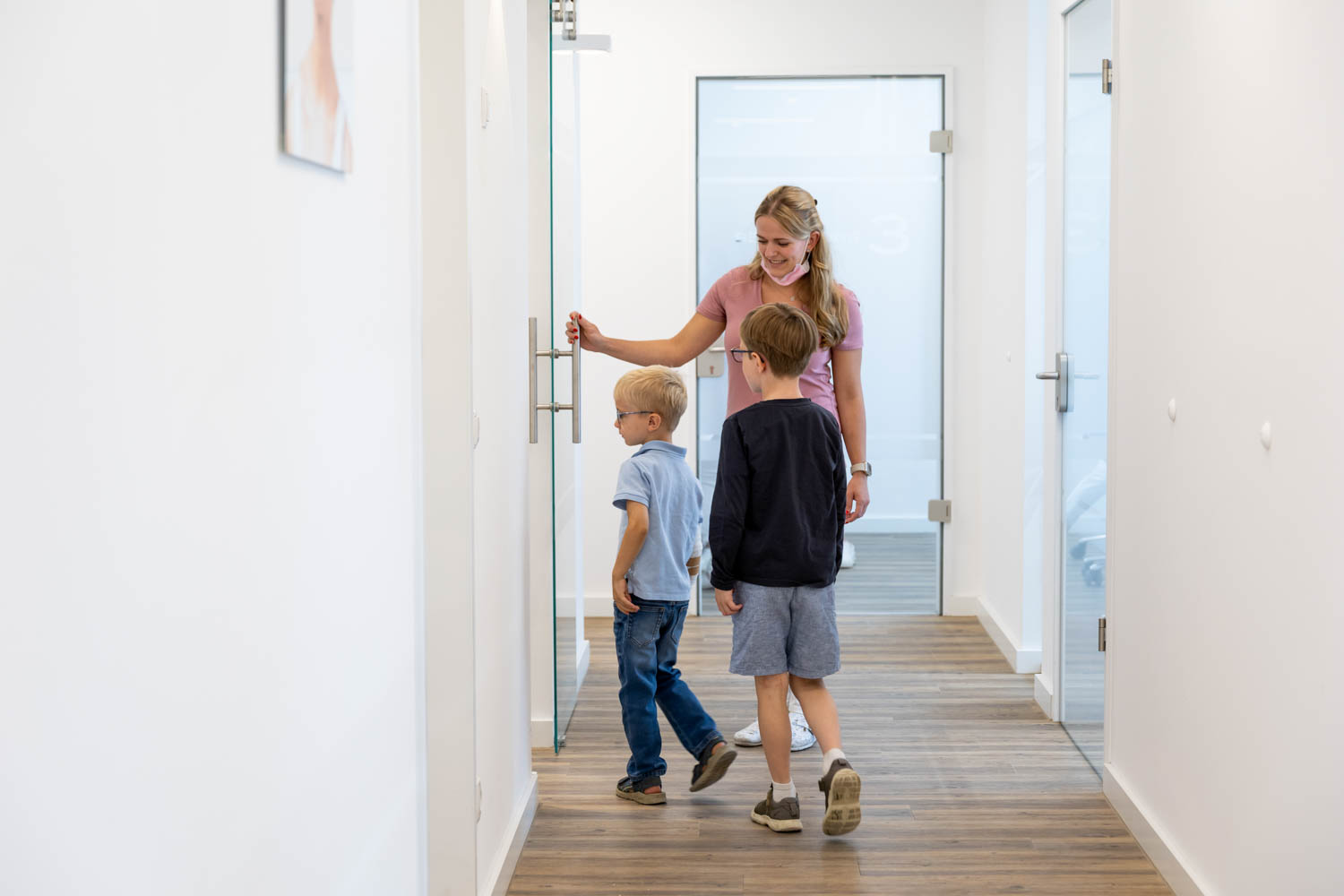 Mitarbeiterin begleitet zwei Kinder freundlich durch den Praxisflur ins Behandlungszimmer der Kieferorthopädie Seelscheid in Neunkirchen-Seelscheid.