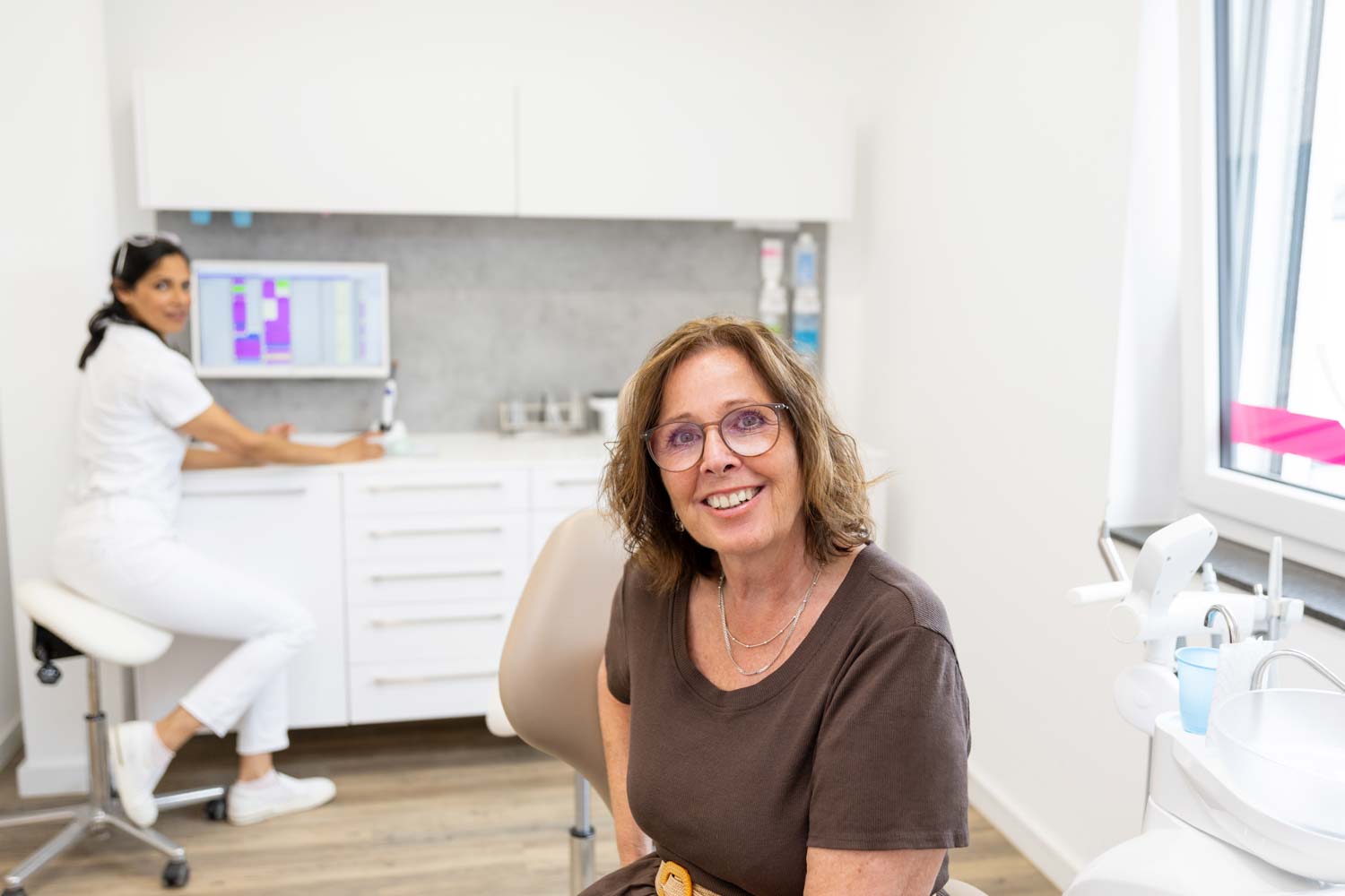 Lächelnde erwachsene Patientin (Best Ager) im Behandlungsstuhl der Kieferorthopädie Seelscheid in Neunkirchen-Seelscheid, im Hintergrund Kieferorthopädin Dr. Garcia.