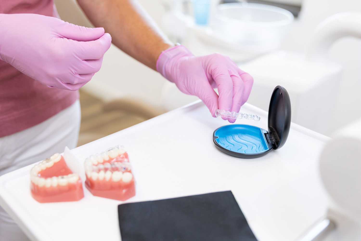 Hände in rosa Handschuhen legen eine transparente Invisalign-Aligner-Schiene in eine Etui-Dose in der Kieferorthopädie Seelscheid in Neunkirchen-Seelscheid.