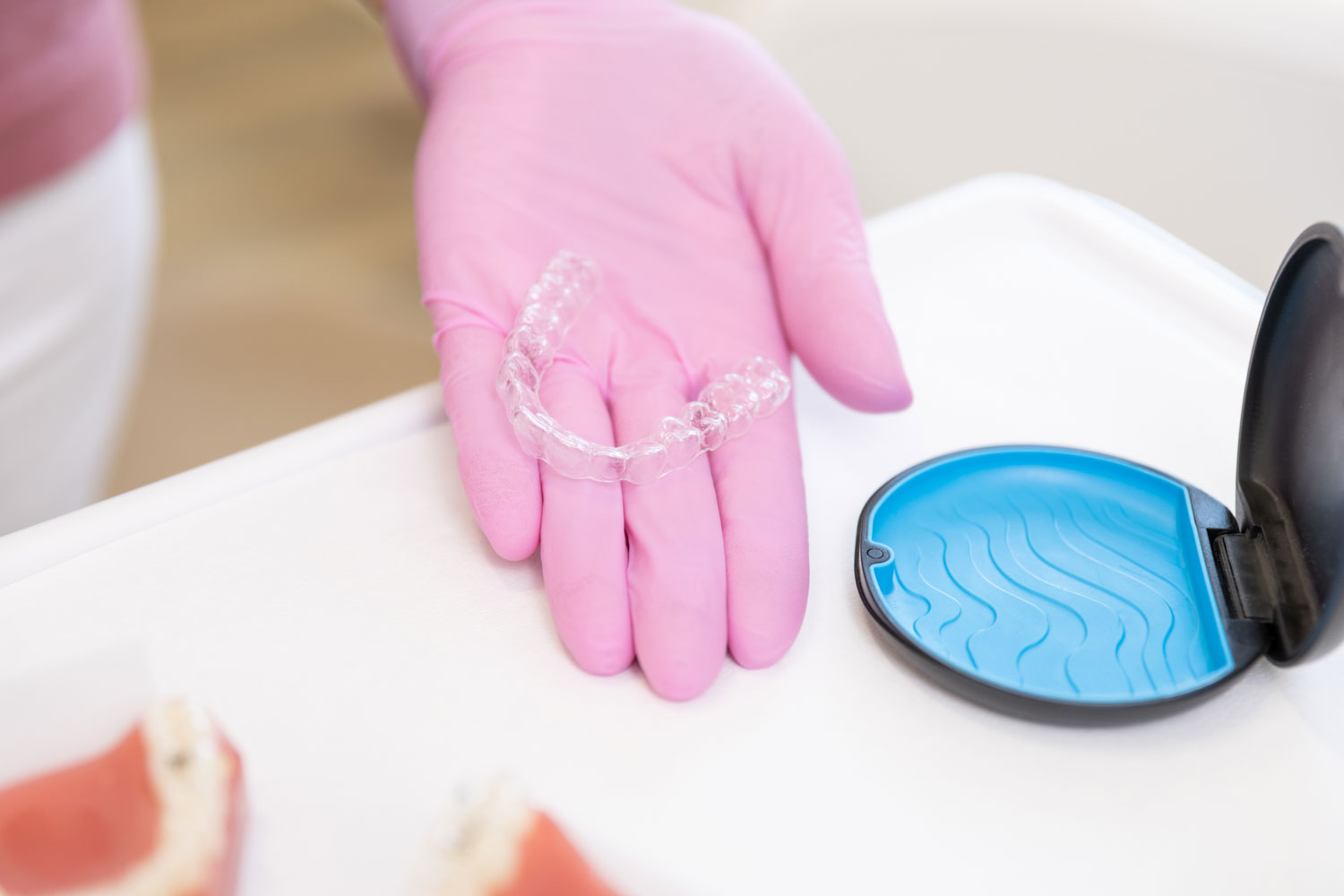 Eine Hand in rosa Einwegschuh präsentiert eine transparente Invisalign-Aligner-Schiene neben einer blau-schwarzen Aufbewahrungsdose in der Praxis Kieferorthopädie Seelscheid in Neunkirchen-Seelscheid.