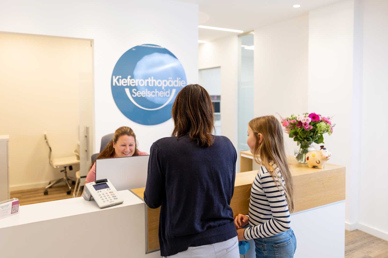 Eine Mutter und ihre Tochter stehen am Empfangstresen und werden freundlich von einer Mitarbeiterin der Praxis Kieferorthopädie Seelscheid in Neunkirchen-Seelscheid begrüßt.