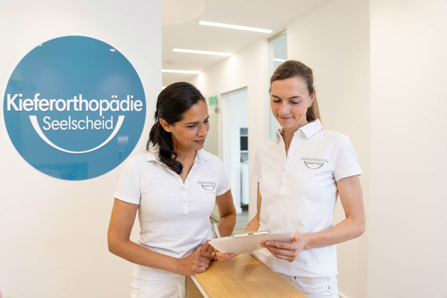 Dr. Garcia und Dr. Schumacher schauen gemeinsam auf Unterlagen auf einem Klemmbrett am Empfang der Praxis Kieferorthopädie Seelscheid in Neunkirchen-Seelscheid.