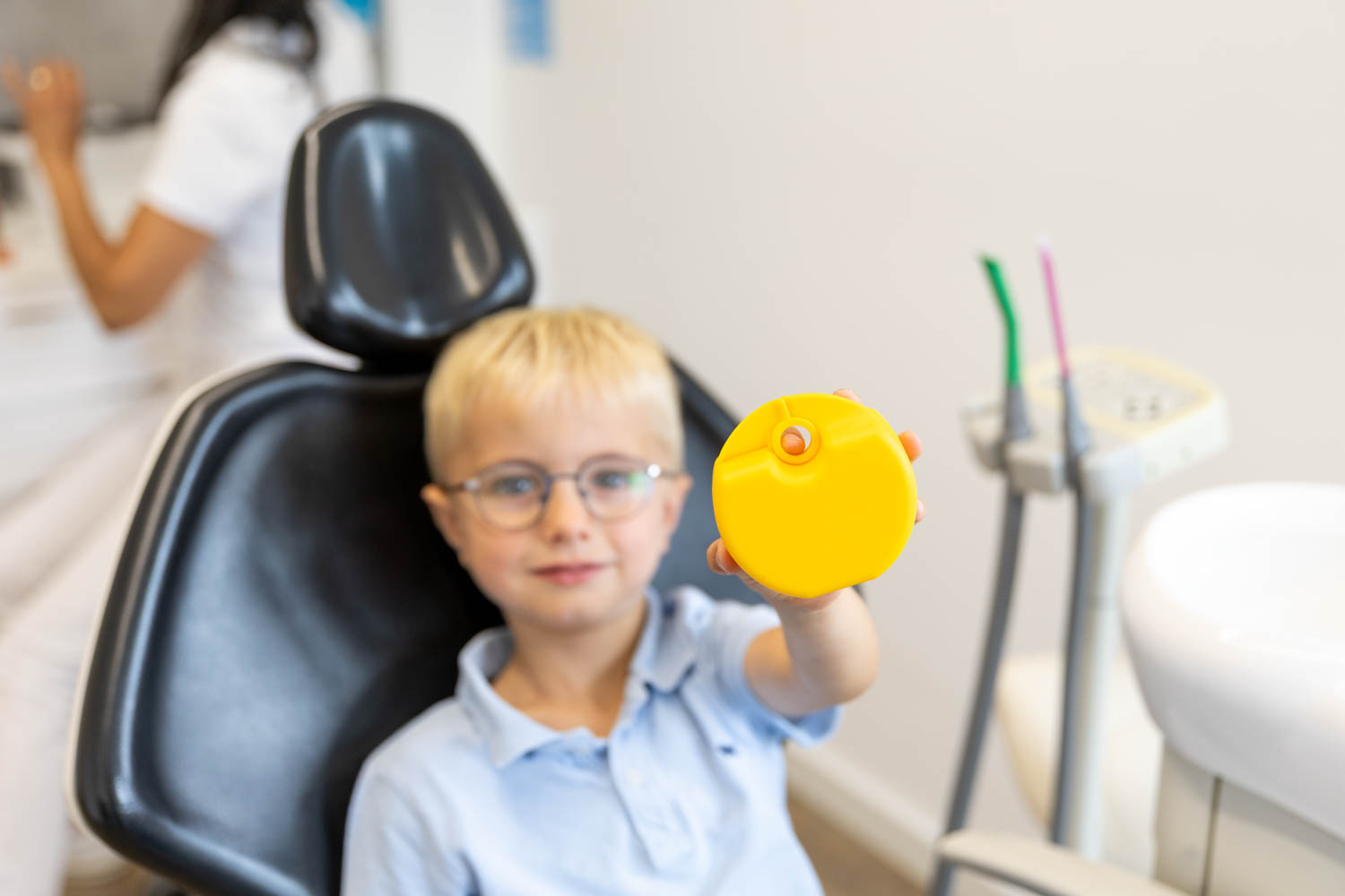 Ein kleiner Junge mit Brille hält auf dem Behandlungsstuhl der Kieferorthopädie Seelscheid stolz seine gelbe Dose für eine Zahnspange in die Kamera.