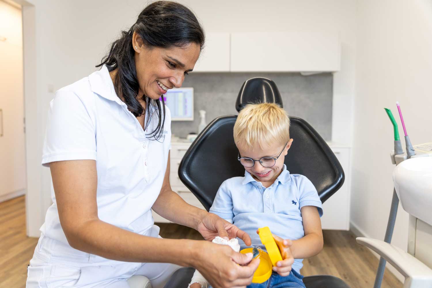 Kieferorthopädin Dr. Garcia erklärt einem Jungen mit Brille auf dem Behandlungsstuhl eine gelbe Zahnspangendose in der Praxis Kieferorthopädie Seelscheid in Neunkirchen-Seelscheid.