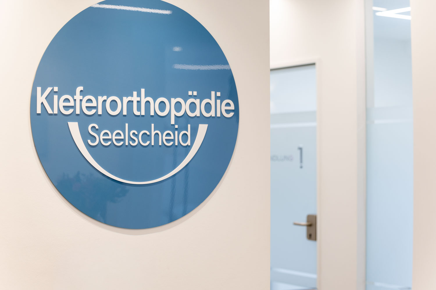 Alternativtext (Alt-Text) Blaues Praxislogo der Kieferorthopädie Seelscheid an der Flurwand vor dem Behandlungszimmer 1 in Neunkirchen-Seelscheid.