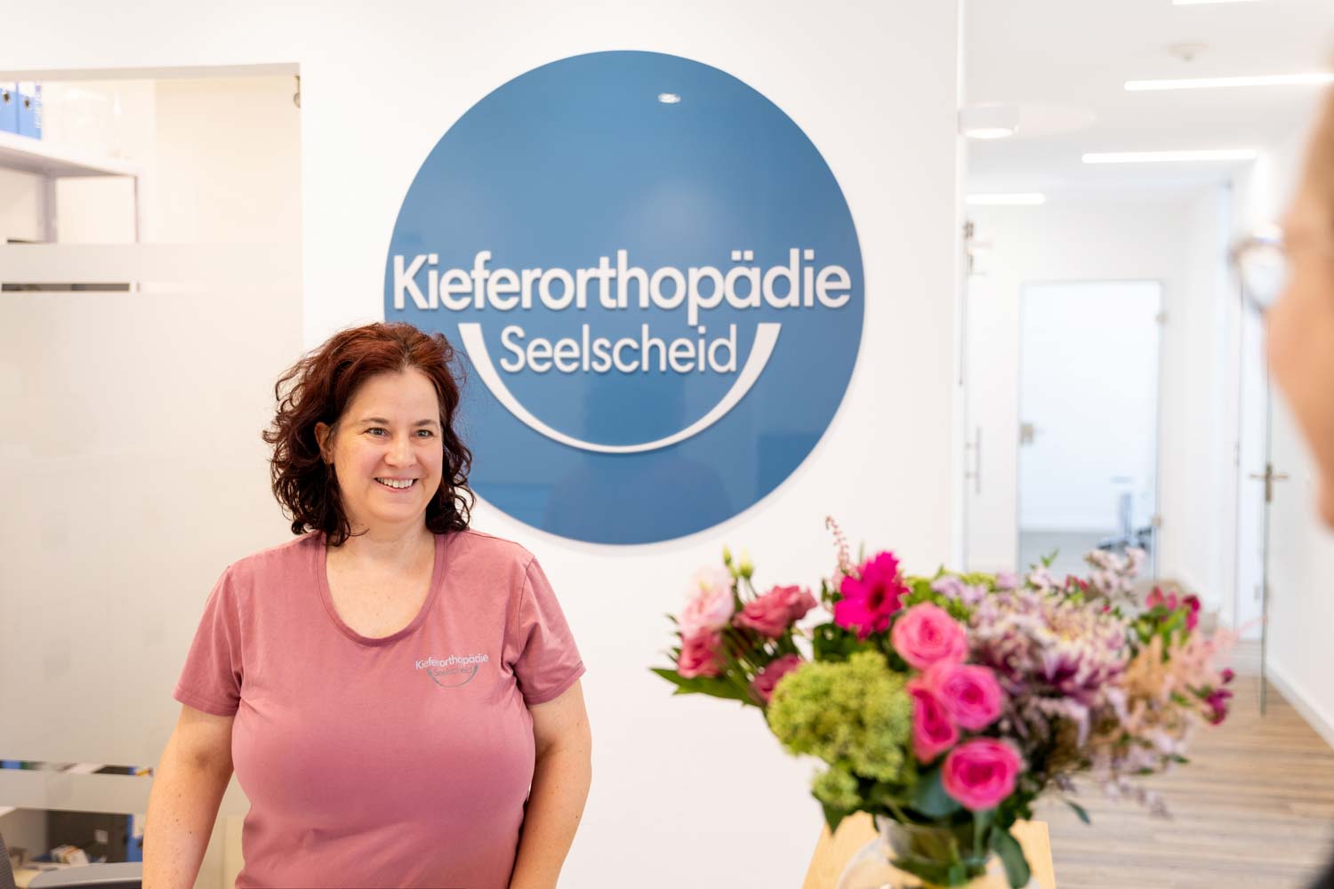 Lächelnde Mitarbeiterin im Praxis-Outfit vor dem Wandlogo am Empfang der Kieferorthopädie Seelscheid in Neunkirchen-Seelscheid.